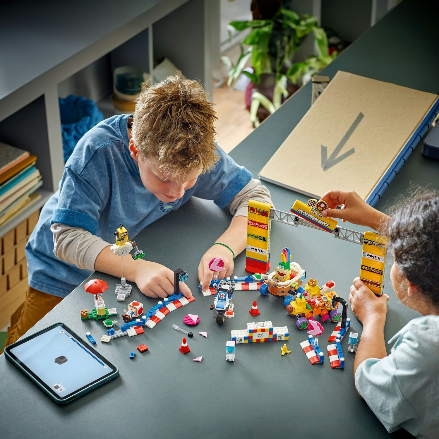 Due bambini giocano con set Lego di Mario Kart su un tavolo grigio. Sono visibili macchine, personaggi, piste e un tablet Lego.
