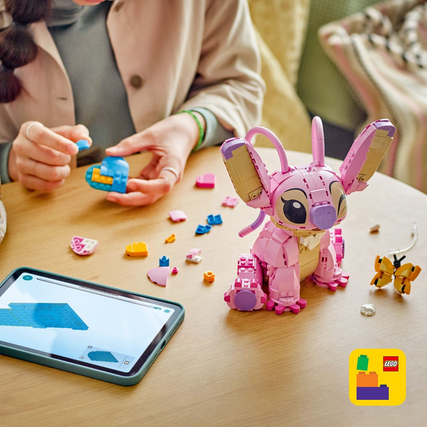 Una persona assembla un personaggio Lego rosa con un tablet che mostra le istruzioni. Pezzi Lego, inclusi cuori, sono su un tavolo di legno.