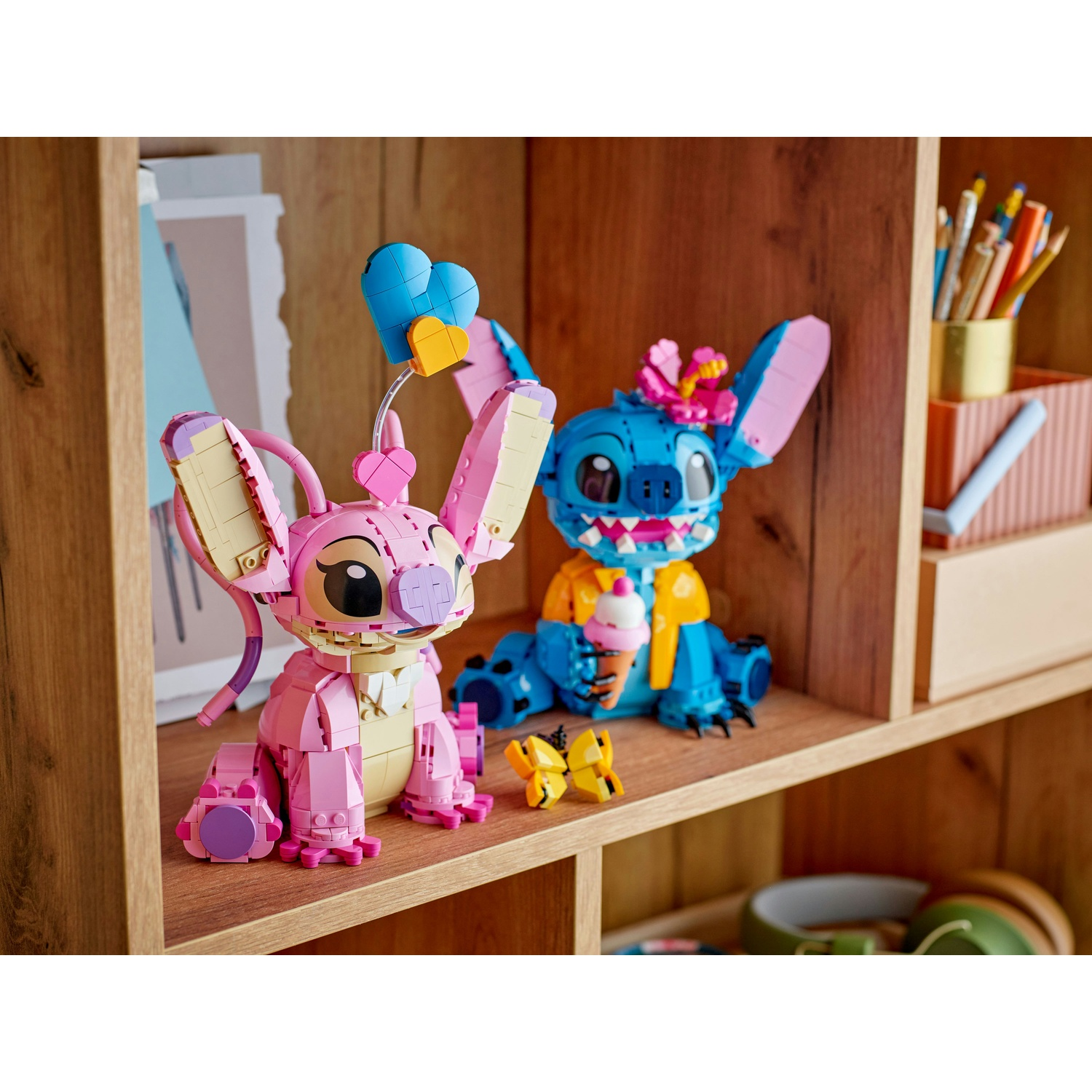 Due personaggi Lego di Stitch e Angel su uno scaffale. Angel è rosa, tiene dei cuori. Stitch è blu, tiene un gelato. Farfalle.
