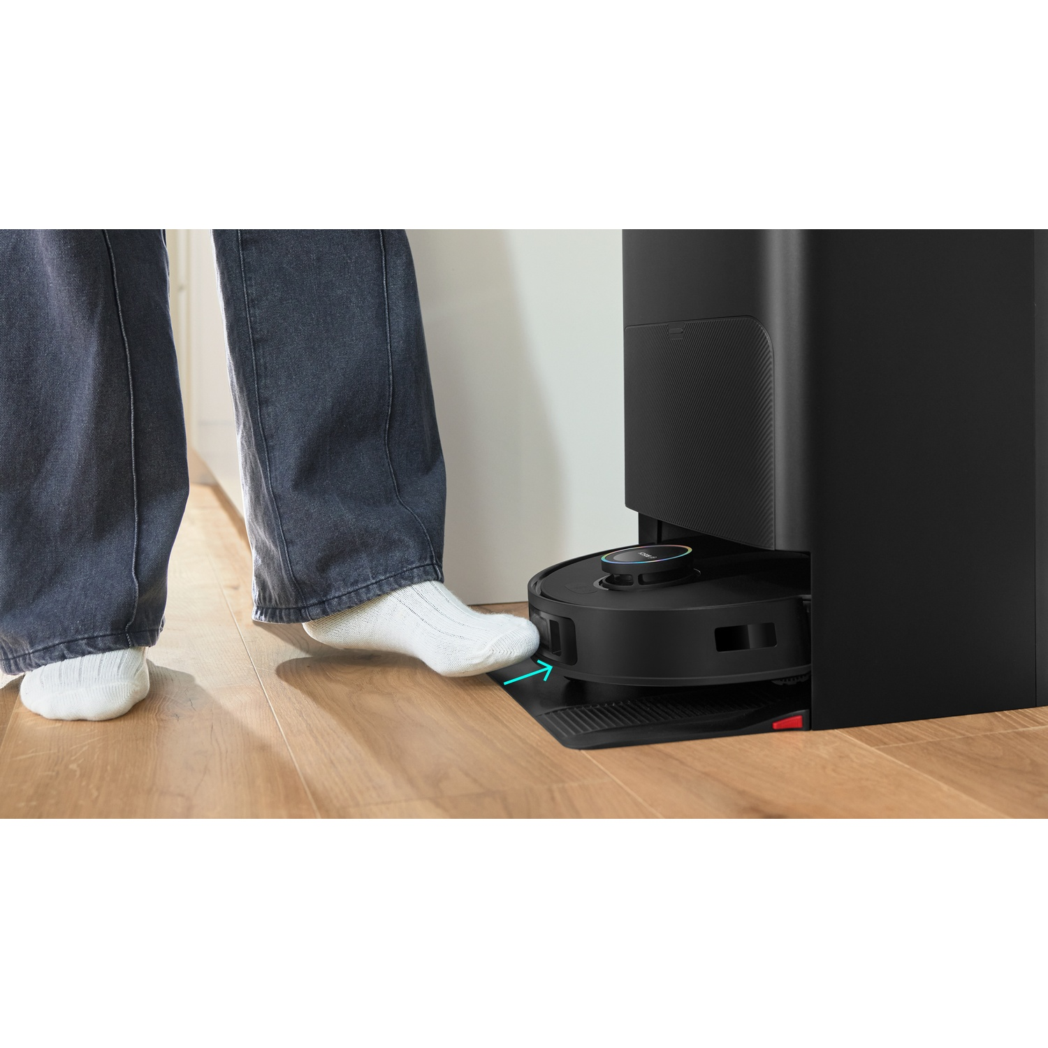 Een voet in witte sok raakt een robotstofzuiger aan. De stofzuiger is zwart en staat op een houten vloer, naast een zwart laadstation.