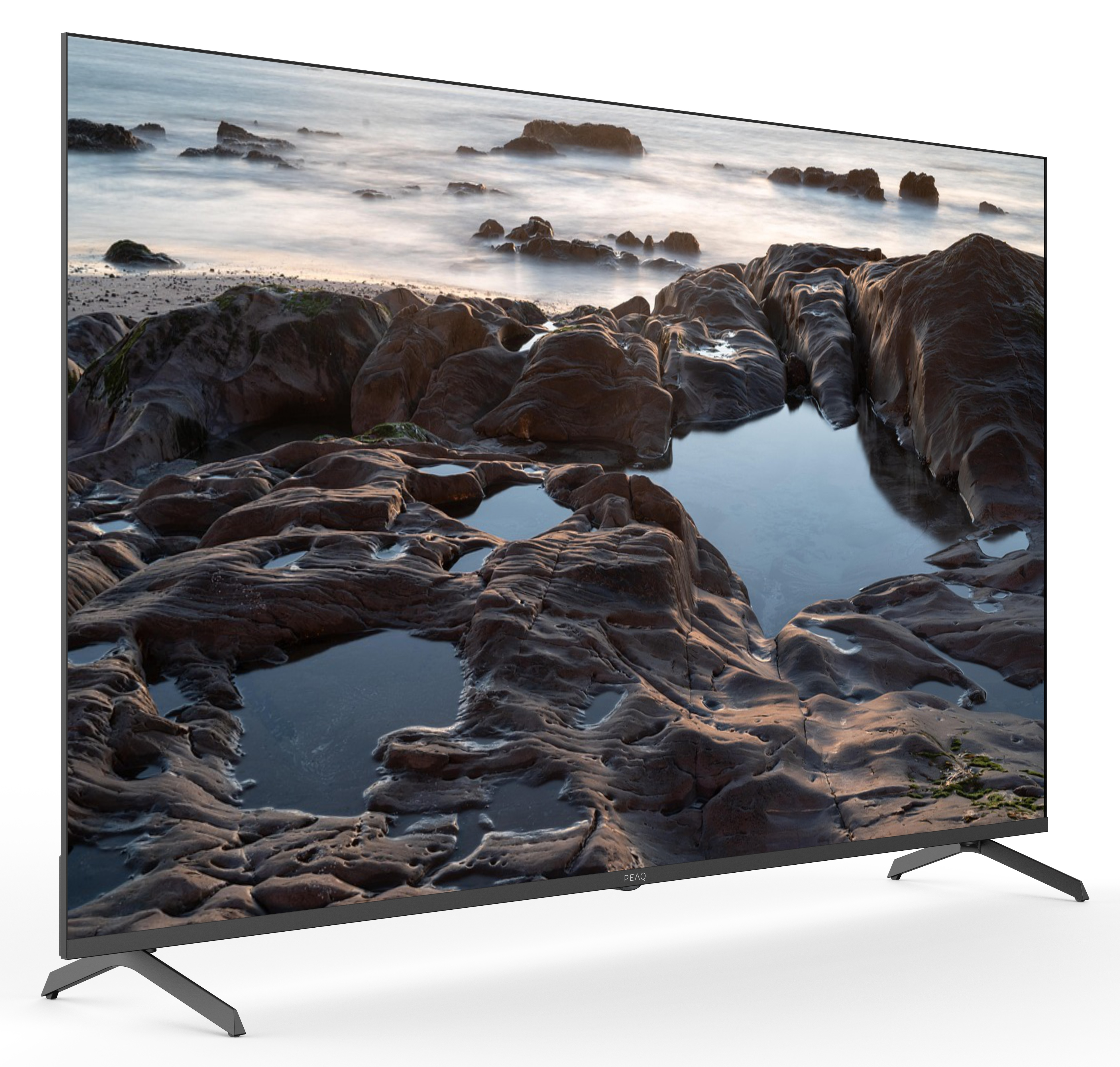 Ekran telewizora wyświetla krajobraz morski, prezentując formacje skalne i wodę. Telewizor ma czarną ramkę, stoi na nóżkach i jest na białej powierzchni.