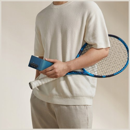 Osoba trzyma niebieski telefon i rakietę tenisową. Osoba ma na sobie kremową koszulę i spodnie. Tło jest jasnobeżowe.