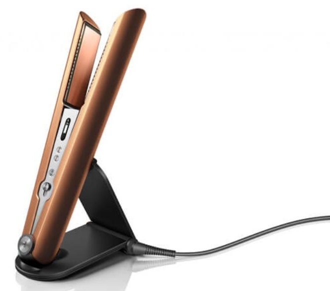 Miedziana prostownica do włosów stoi na czarnym stojaku. Ma podłączony przewód zasilający. Tło jest białe.