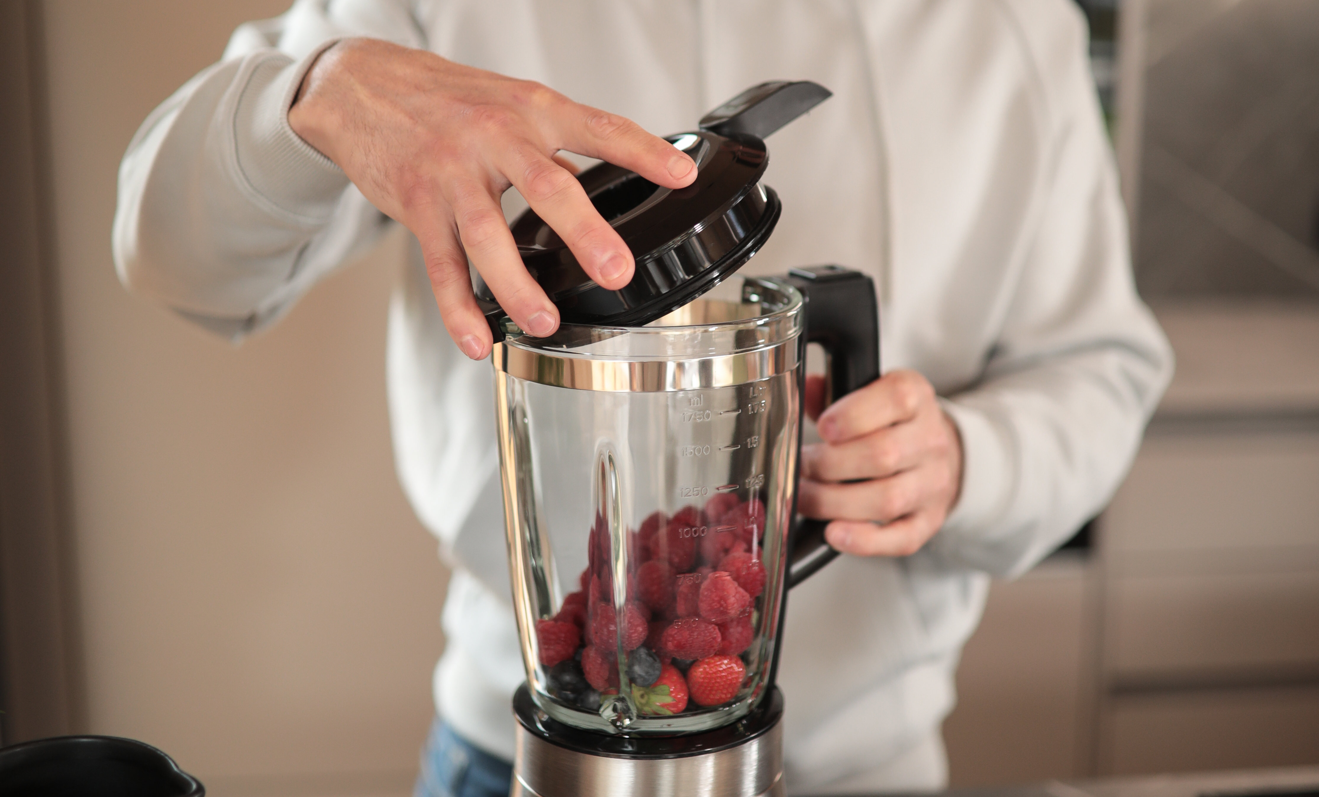 Bir kişi blender kapağını açıyor. Blender meyveler içeriyor. Arka plan bir mutfak ortamı.