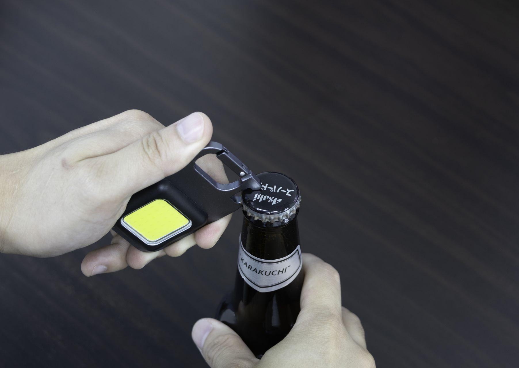 Eller, siyah bir şişe açacağı ile bir bira şişesini açıyor. Kapakta Asahi yazıyor.