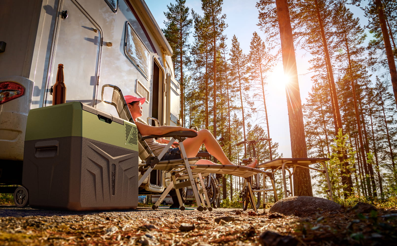 Kobieta odpoczywa na krześle obok kampera. Obok znajduje się lodówka i butelka. Wysokie drzewa i światło słoneczne wypełniają tło.