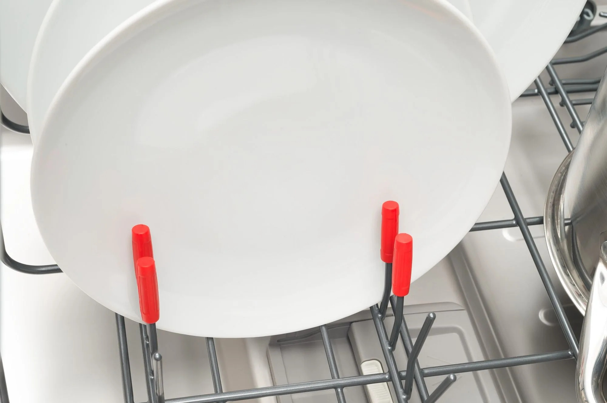 Białe talerze w zmywarce, wspierane przez czerwone uchwyty. Metalowy garnek po prawej. Scena jest jasna i czysta.