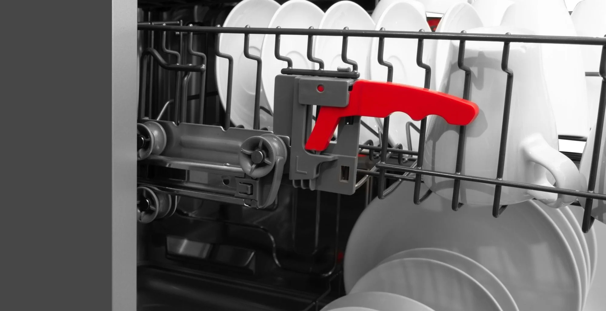 Wnętrze zmywarki. Białe talerze i kubki w stojakach. Czerwony uchwyt na szarym mechanizmie.