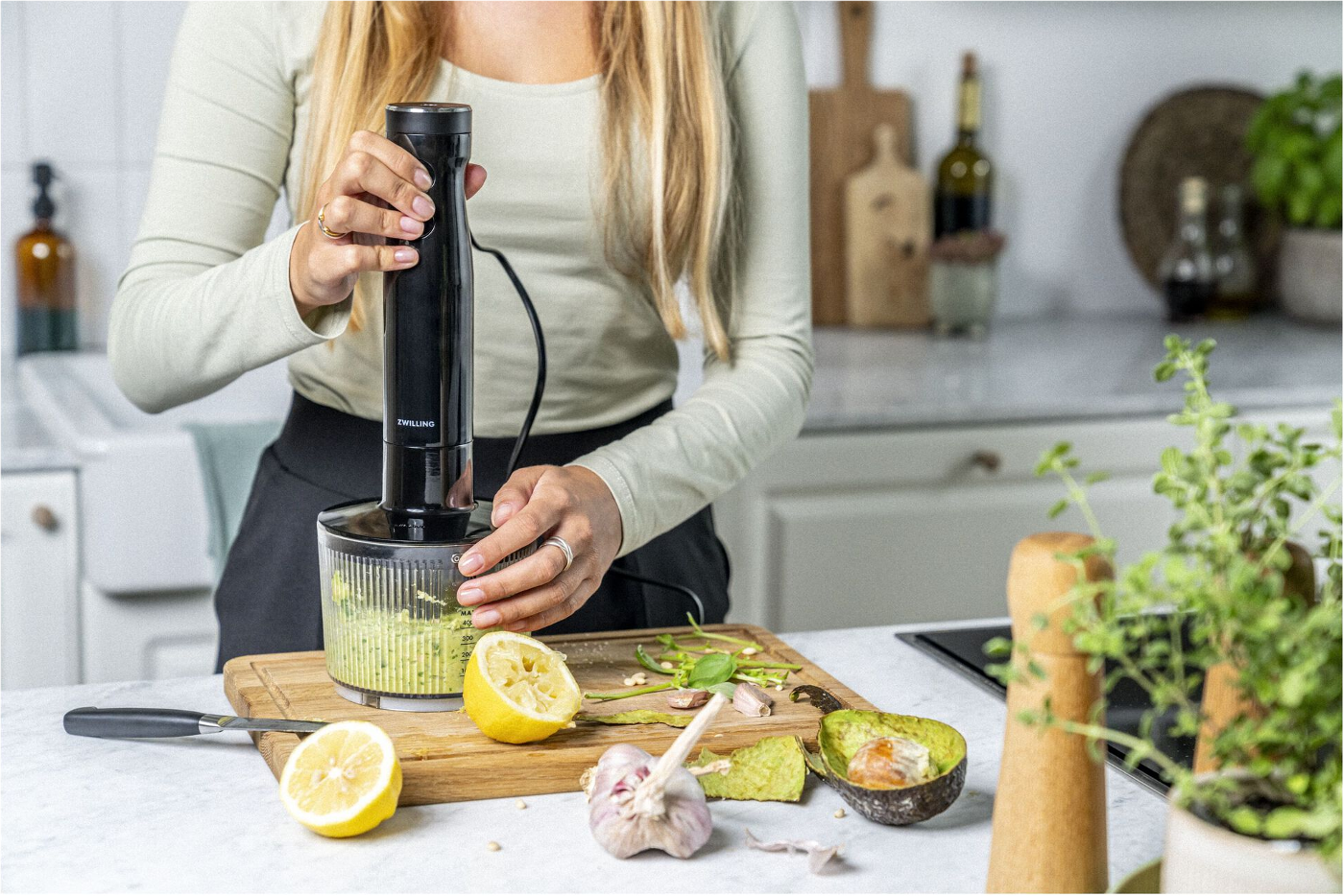 Eine Frau benutzt einen Mixer in einer Küche. Zutaten wie Zitrone, Knoblauch und Avocado liegen auf einem Schneidebrett.