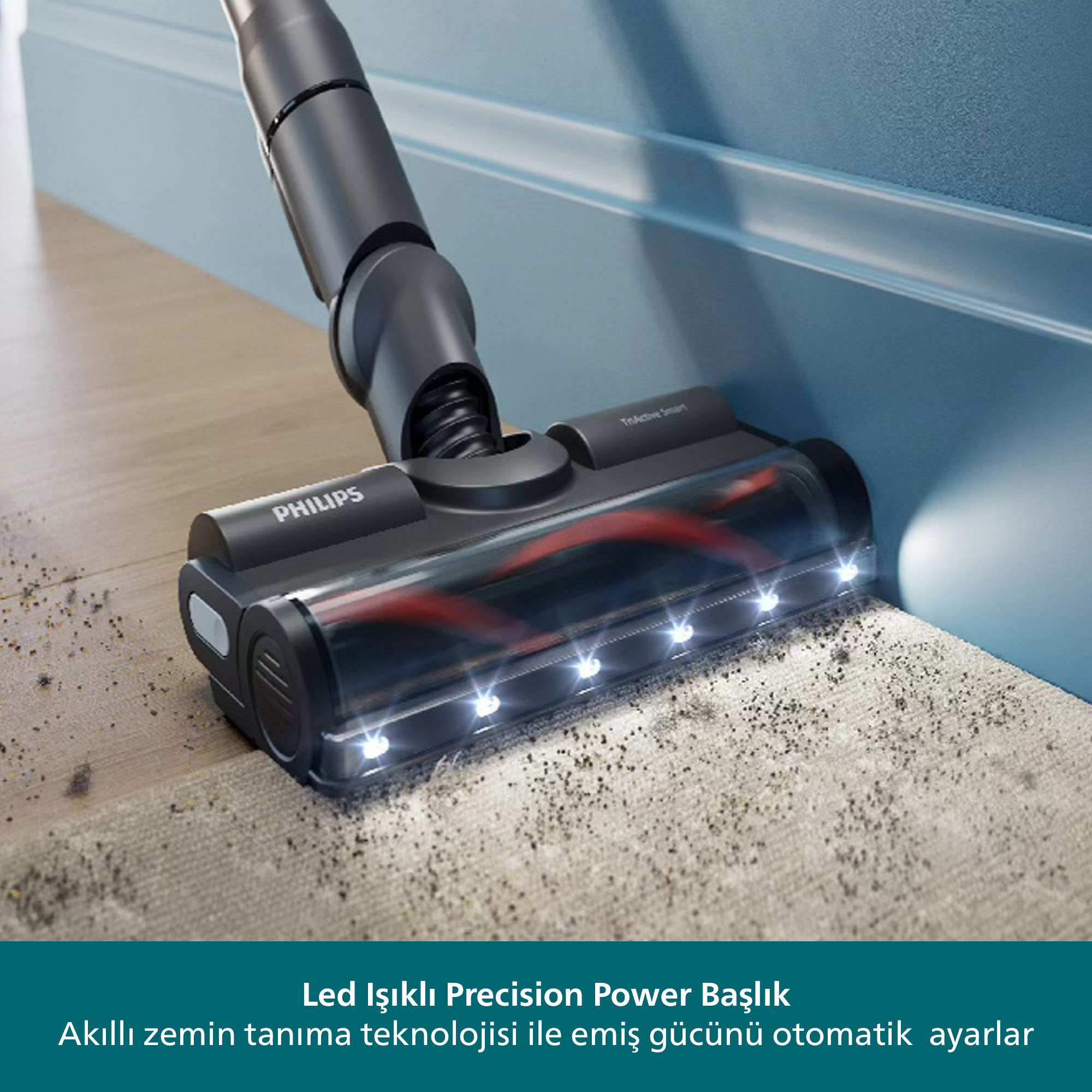 Siyah bir Philips elektrikli süpürge başlığı, mavi bir duvarın yanında, halı üzerinde duruyor. Halıda döküntüler görülüyor.