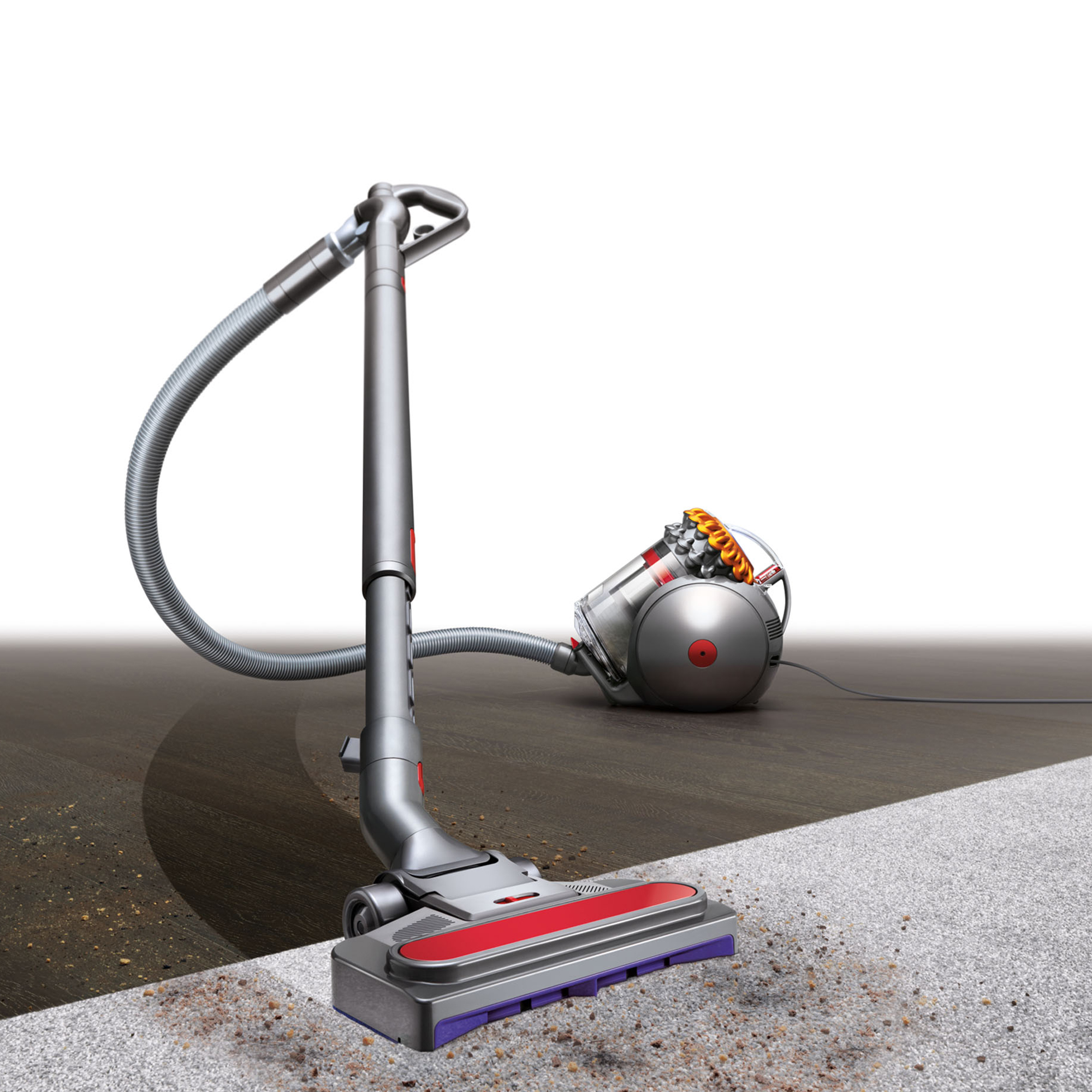 İki renkli bir zeminde gümüş ve kırmızı bir Dyson elektrikli süpürgesi. Halıda kir görülüyor. Elektrikli süpürge ahşap zeminde duruyor.