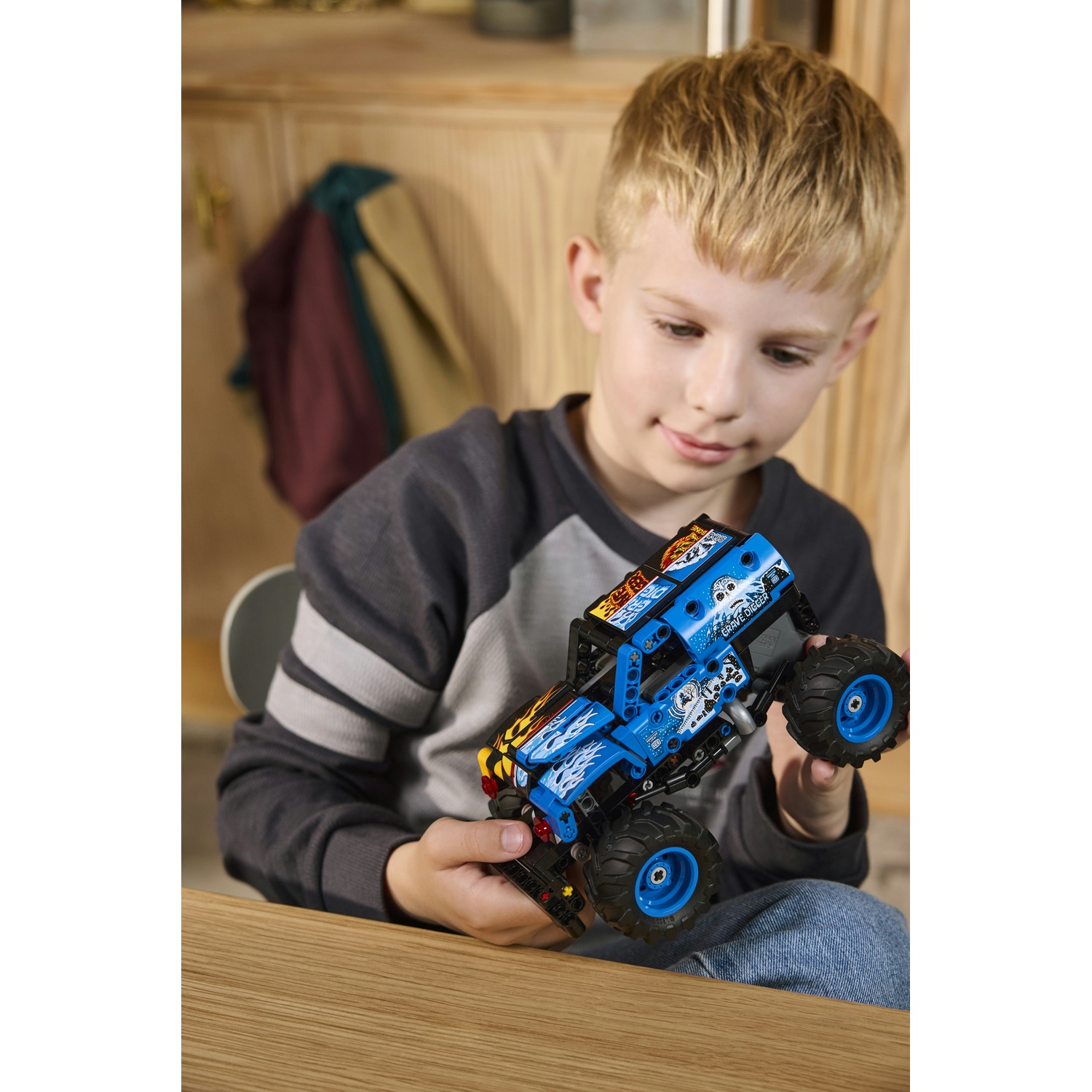 Un ragazzino tiene in mano un camion mostro giocattolo blu. Lo guarda sorridendo. Lo sfondo è un mobile in legno e un cappotto.
