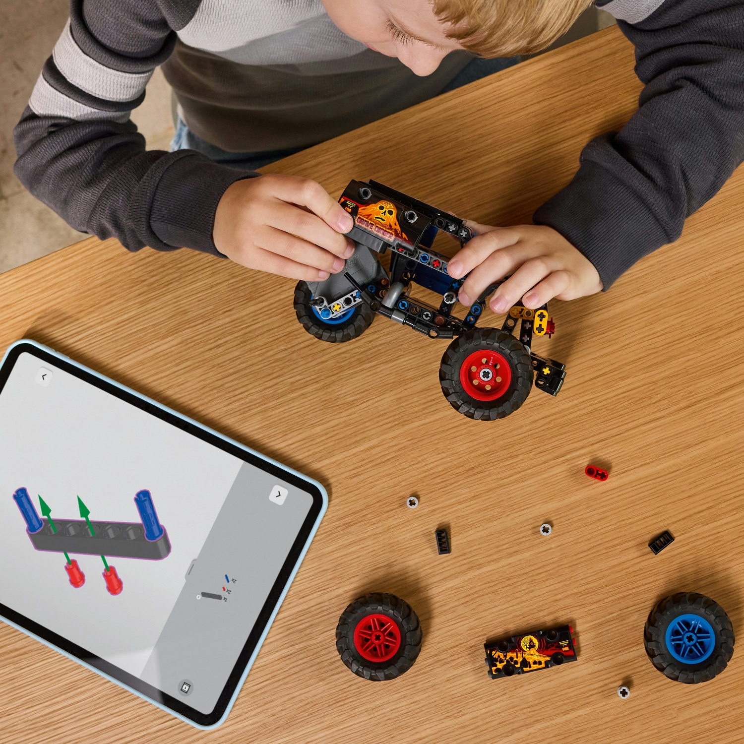 Un bambino sta costruendo un'auto giocattolo con parti nere e rosse. Un iPad mostra le istruzioni. La scena è su un tavolo di legno.