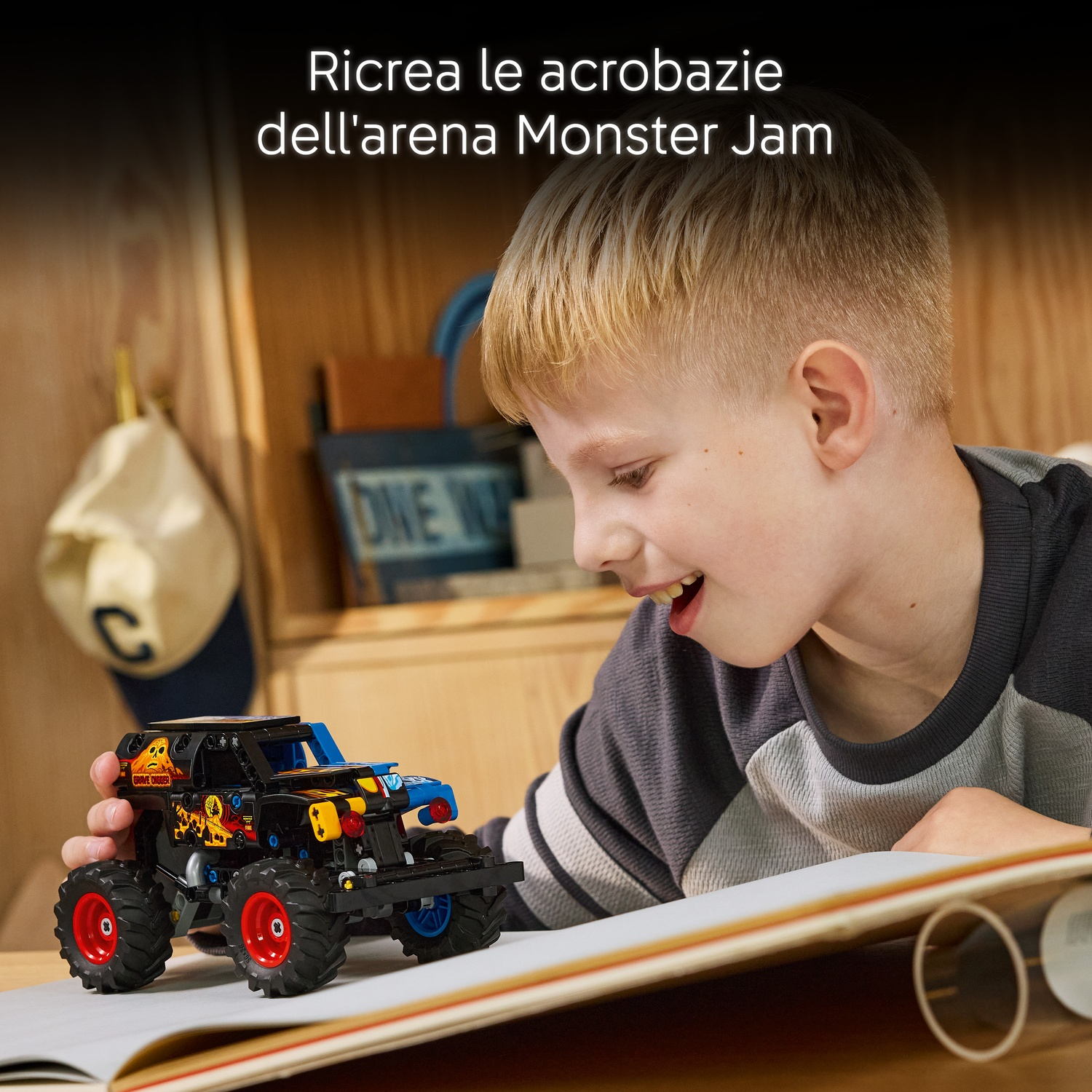 Un ragazzo guarda un monster truck giocattolo su un tavolo. Il camion è nero, blu e giallo con ruote rosse. Un berretto è appeso sullo sfondo.