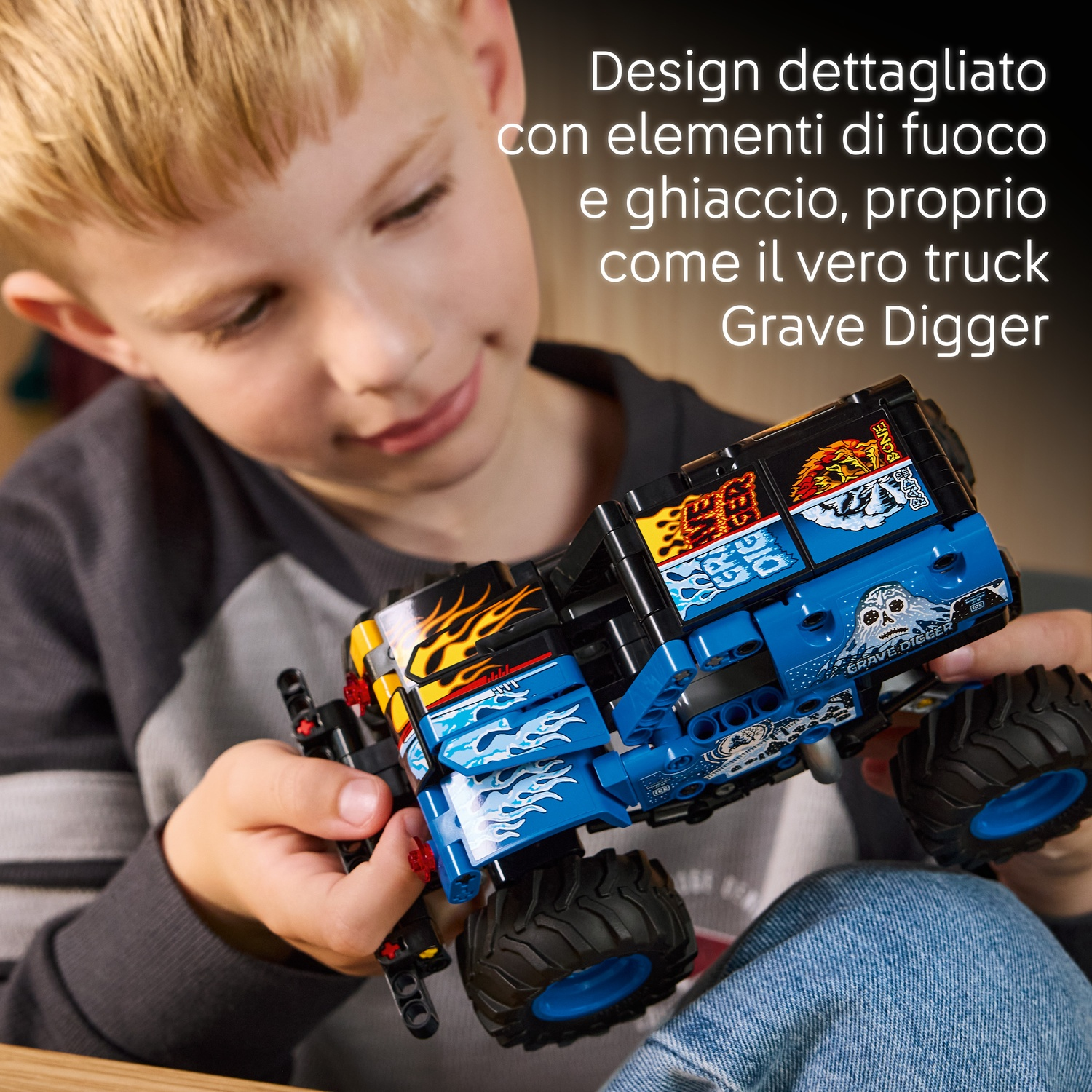 Un bambino tiene in mano un camion giocattolo Grave Digger blu e nero. Il camion ha dettagli di fiamme e ghiaccio. Il bambino indossa una maglia grigia e jeans.