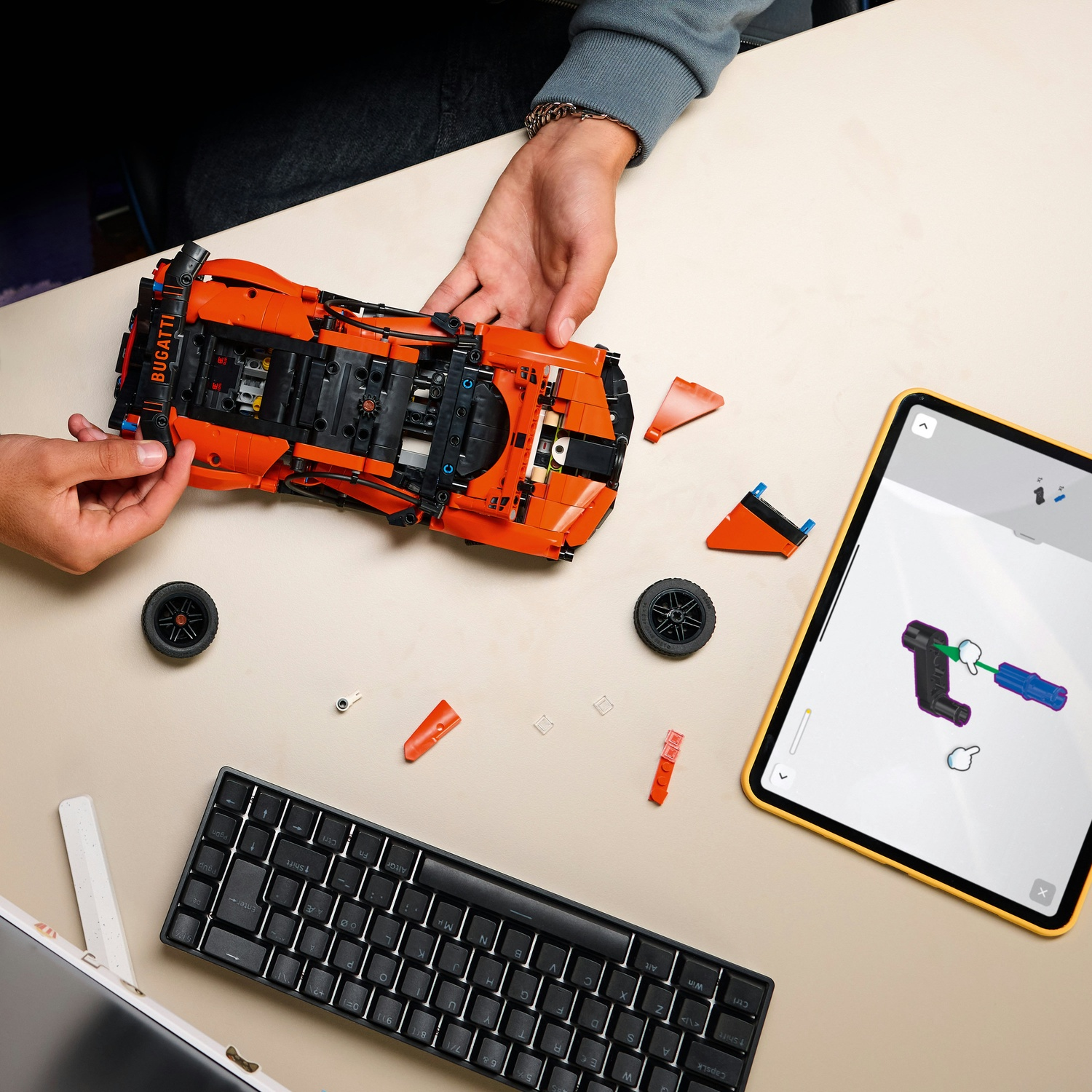 Mani che assemblano un'auto Lego Bugatti arancione. Pezzi, ruote, tastiera e tablet con istruzioni su superficie beige.