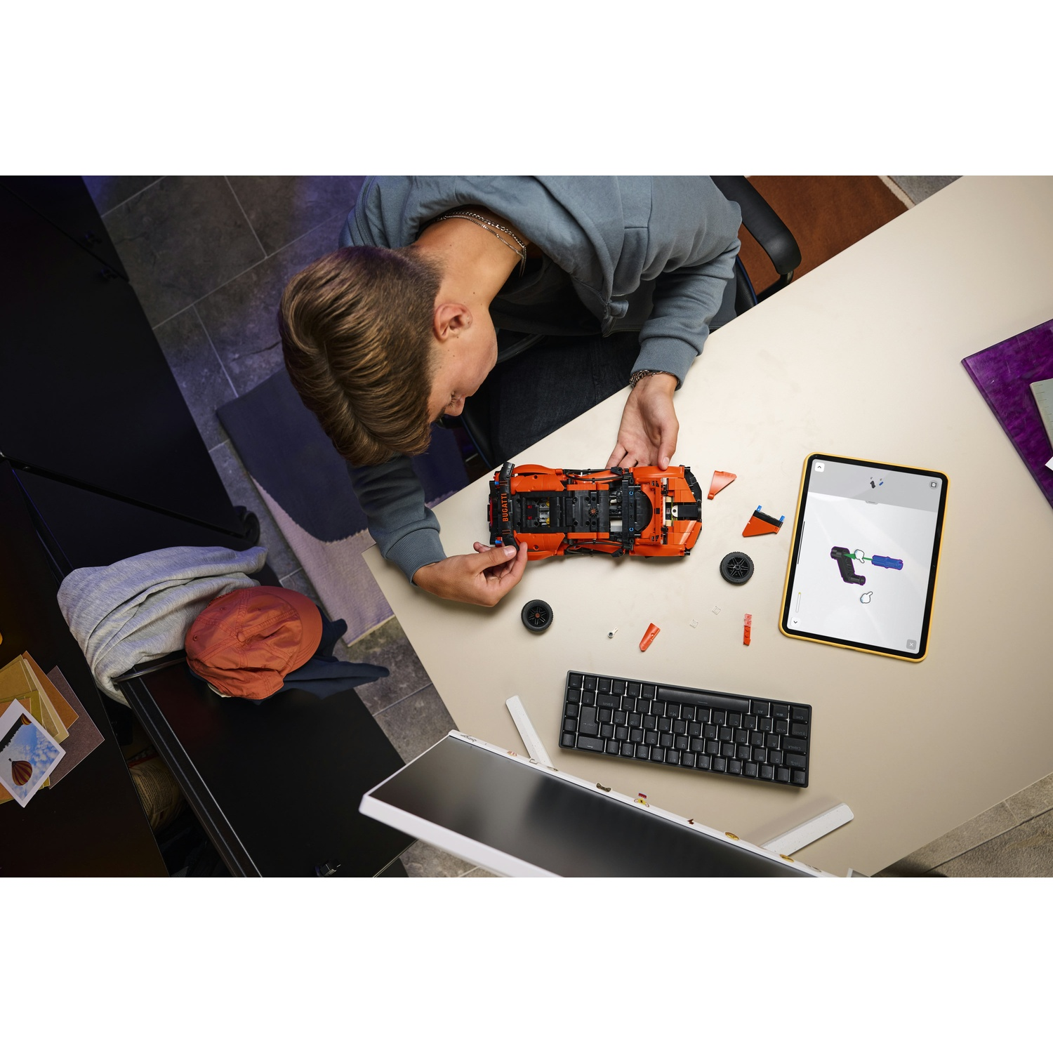 Una persona costruisce un'auto Lego arancione su un tavolo. Parti e un tablet sono vicini. Una tastiera e un monitor sono nella scena.