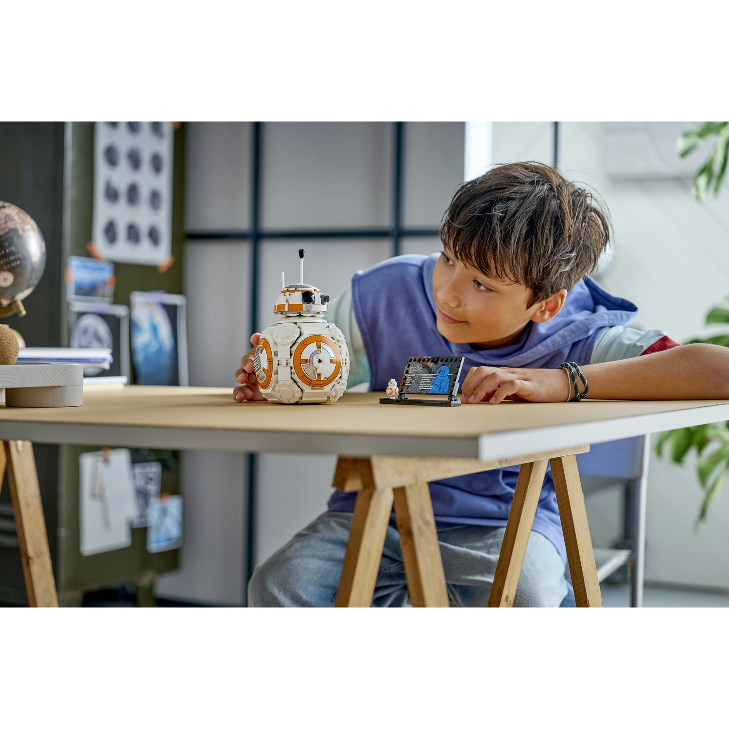 Un ragazzo guarda un droide Lego BB-8 e un piccolo set Lego. Il BB-8 è bianco e arancione. Il ragazzo indossa una felpa blu.