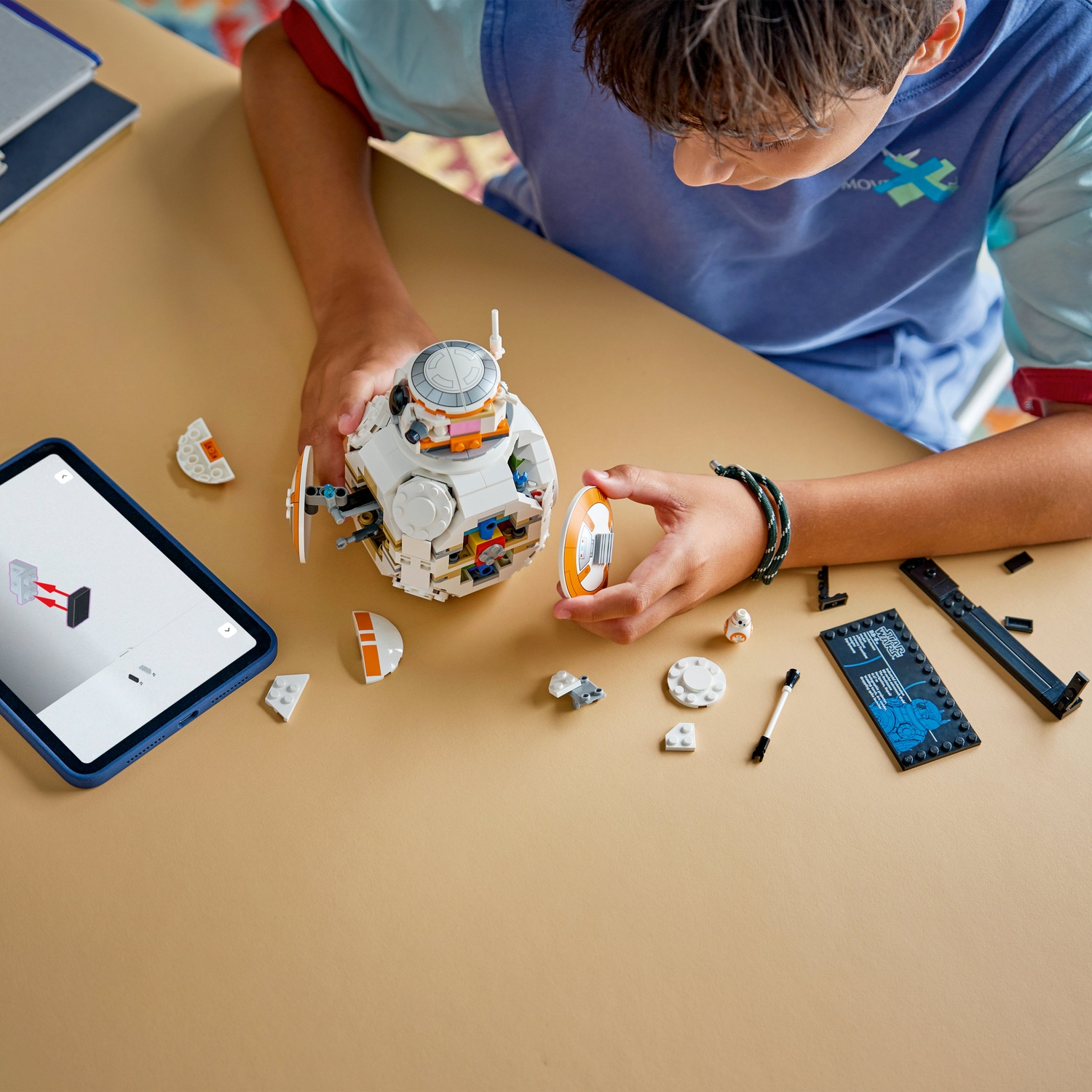 Una persona assembla un droide BB-8 Lego, con pezzi e istruzioni su un tablet. Il droide è bianco e arancione, su una superficie beige.