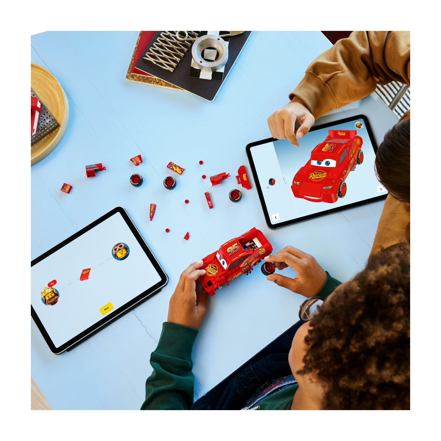 I bambini assemblano un'auto giocattolo rossa, Saetta McQueen, usando parti e tablet con istruzioni su un tavolo azzurro.