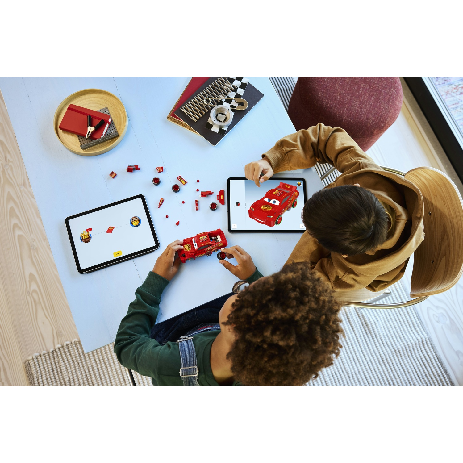 Due bambini costruiscono un'auto da corsa giocattolo rossa, seguendo le istruzioni sui tablet. Parti e accessori sparsi sul tavolo.