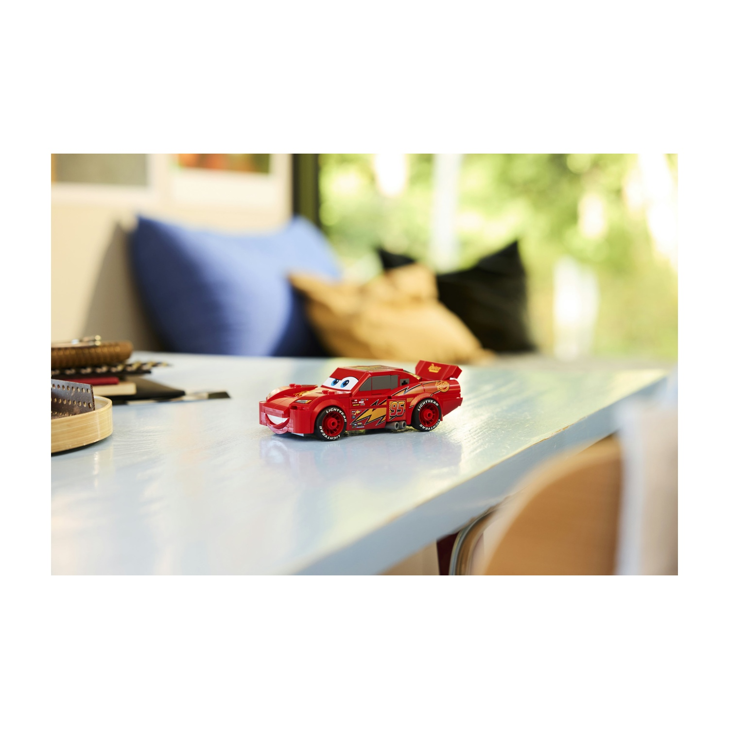 Un'auto giocattolo rossa, Saetta McQueen, è su un tavolo azzurro. Lo sfondo include una finestra, un cuscino blu e vegetazione sfocata.