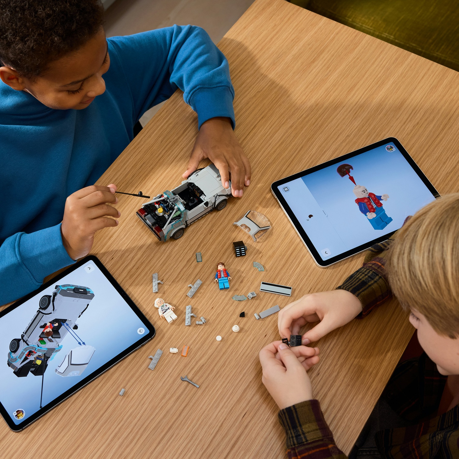 Due bambini costruiscono un'auto Lego, seguendo le istruzioni sui tablet. Pezzi e figure Lego sono sparsi su un tavolo di legno.