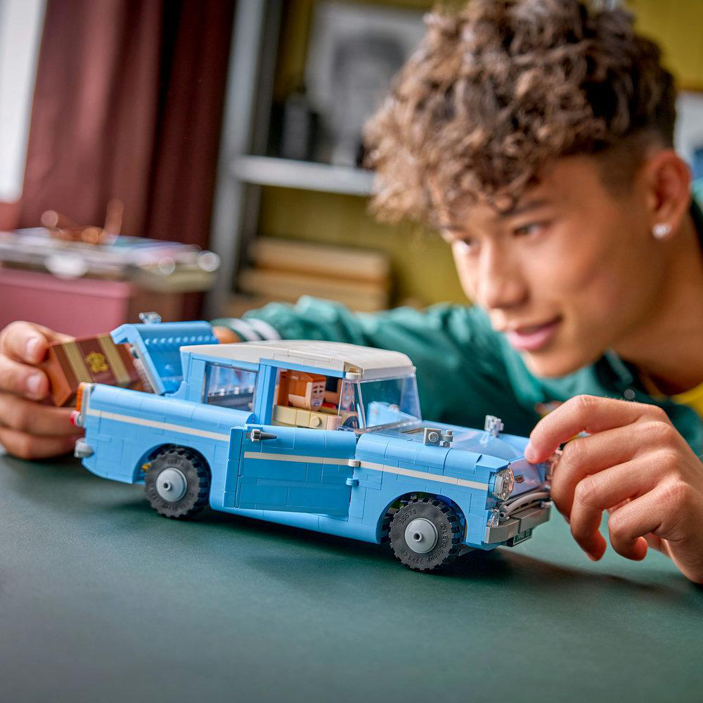 Ein junger Mensch interagiert mit einem hellblauen Lego-Auto mit Koffer. Das Auto steht auf einer grünen Oberfläche, und die Person lächelt.
