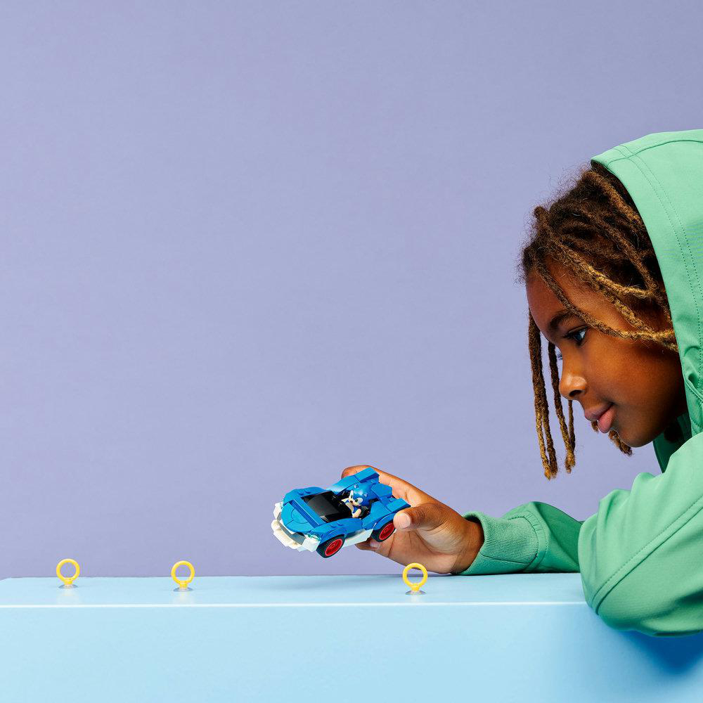 Ein Kind in einem grünen Kapuzenpullover betrachtet ein blaues Spielzeugauto mit einer Sonic-Figur. Drei gelbe Ringe sind vor dem Auto.