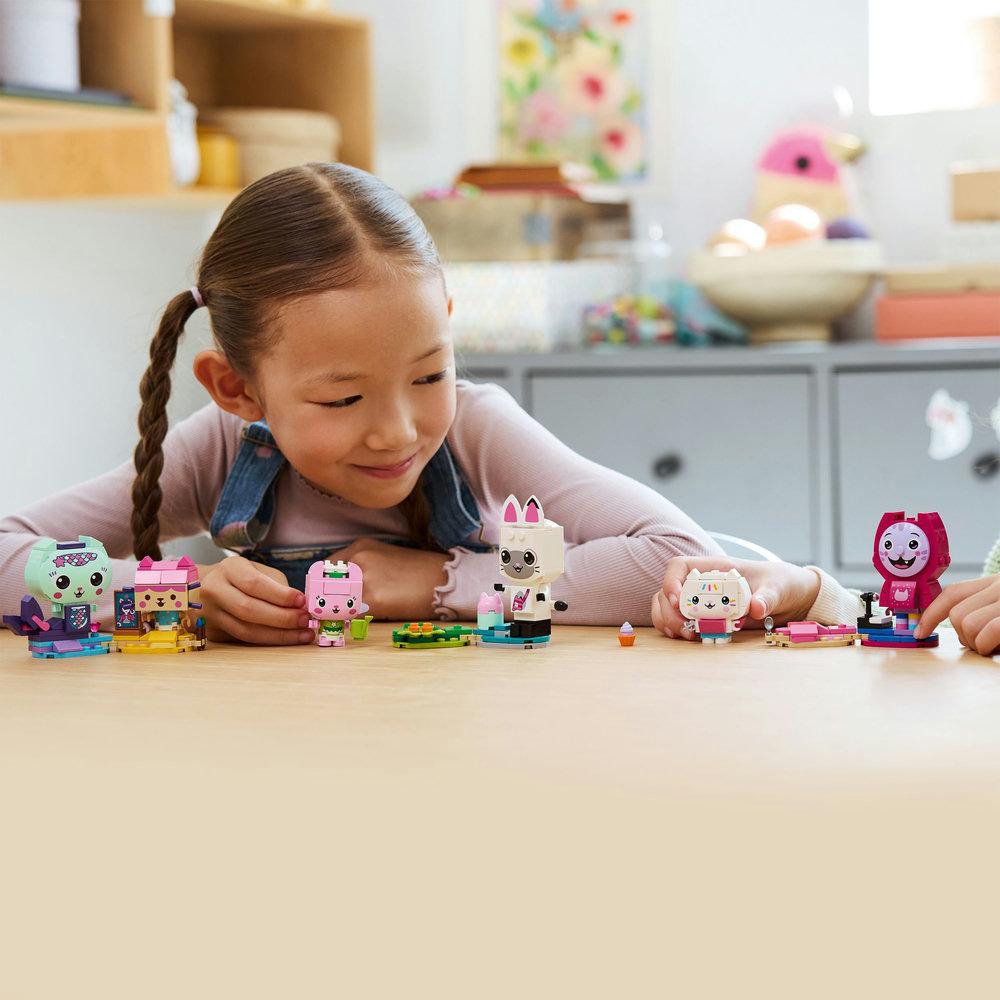 Ein Mädchen lächelt bunte Spielfiguren auf einem Tisch an. Verschiedene Figuren im Katzen-Thema und eine Lama-Figur werden gezeigt.
