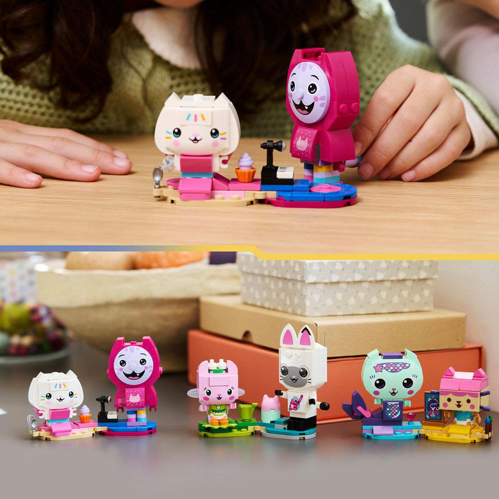 Das Bild zeigt mehrere Spielzeugkatzenfiguren aus Blöcken, einige mit rosa und weißen Farben, die auf einem Tisch angeordnet sind. Der Hintergrund umfasst Boxen.