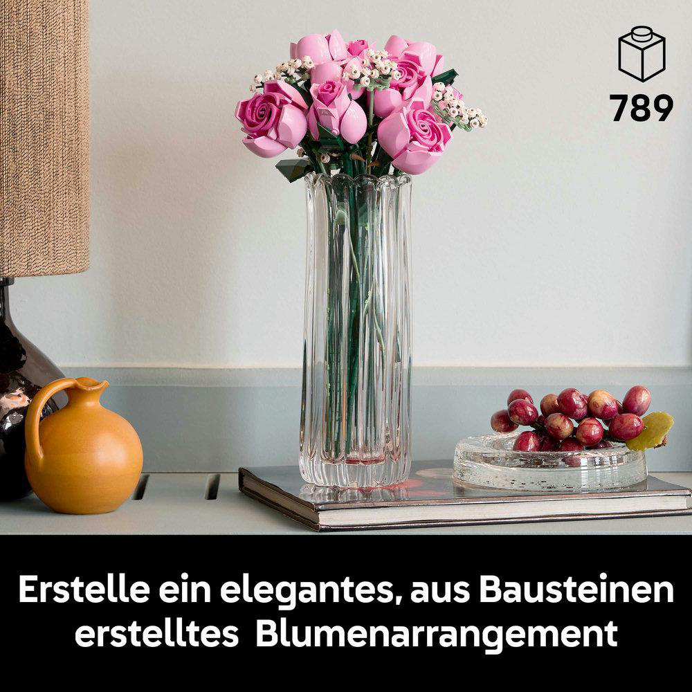Eine Vase mit rosa Rosenblüten. Auf einem Holztisch stehen ein kleiner gelber Krug und eine Traubendekoration. Im Hintergrund eine Lampe.