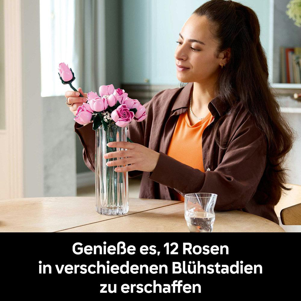 Frau, die rosa Rosen in einer Glasvase arrangiert. Sie trägt ein braunes Hemd über einem orangefarbenen Top. Ein Glas Wasser steht daneben.
