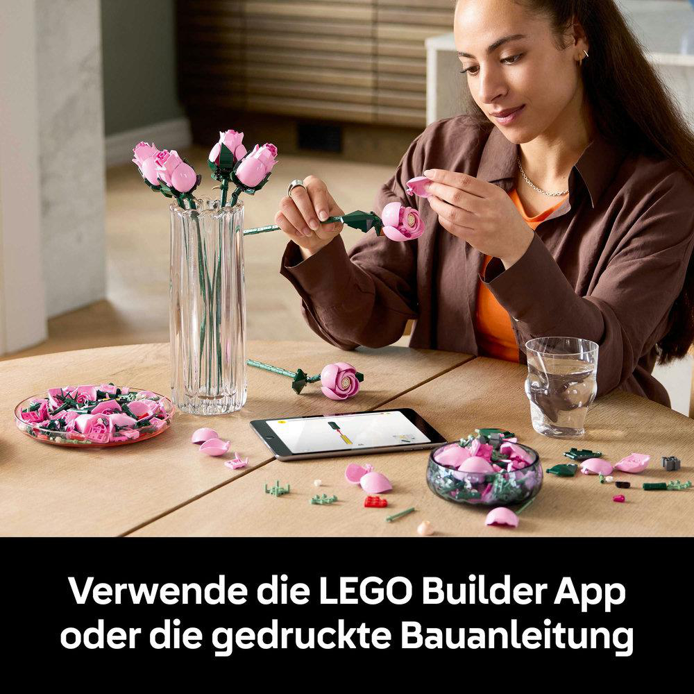 Frau baut rosa LEGO-Rosen zusammen. Auf dem Tisch stehen eine Vase mit Rosen, ein Tablet und ein Glas Wasser.