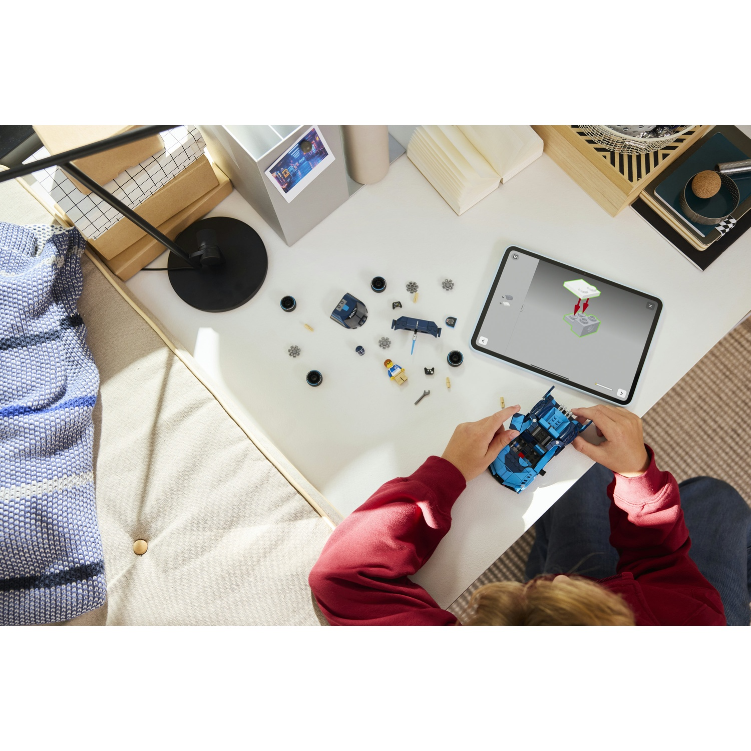 Una persona assembla un'auto Lego blu su un tavolo bianco. Parti e un tablet con istruzioni sono nelle vicinanze. Altri oggetti includono scatole e una lampada.
