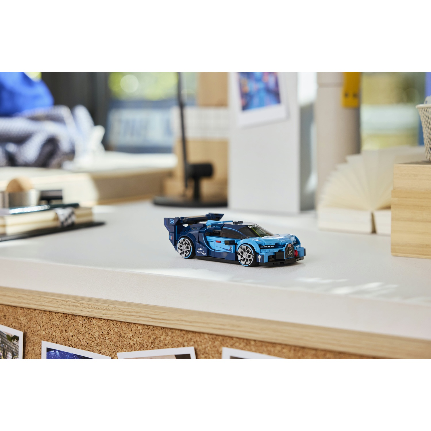 Un'auto giocattolo blu è su una scrivania bianca. Una bacheca con foto è di fronte. Forniture per ufficio e una finestra sullo sfondo.