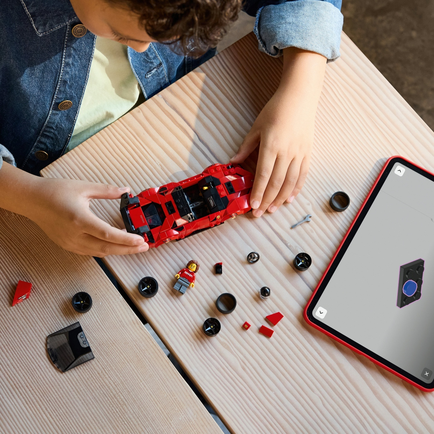 Un bambino assembla un'auto Lego rossa con un tablet, pezzi e una figurina su un tavolo di legno. Il tablet mostra le istruzioni.