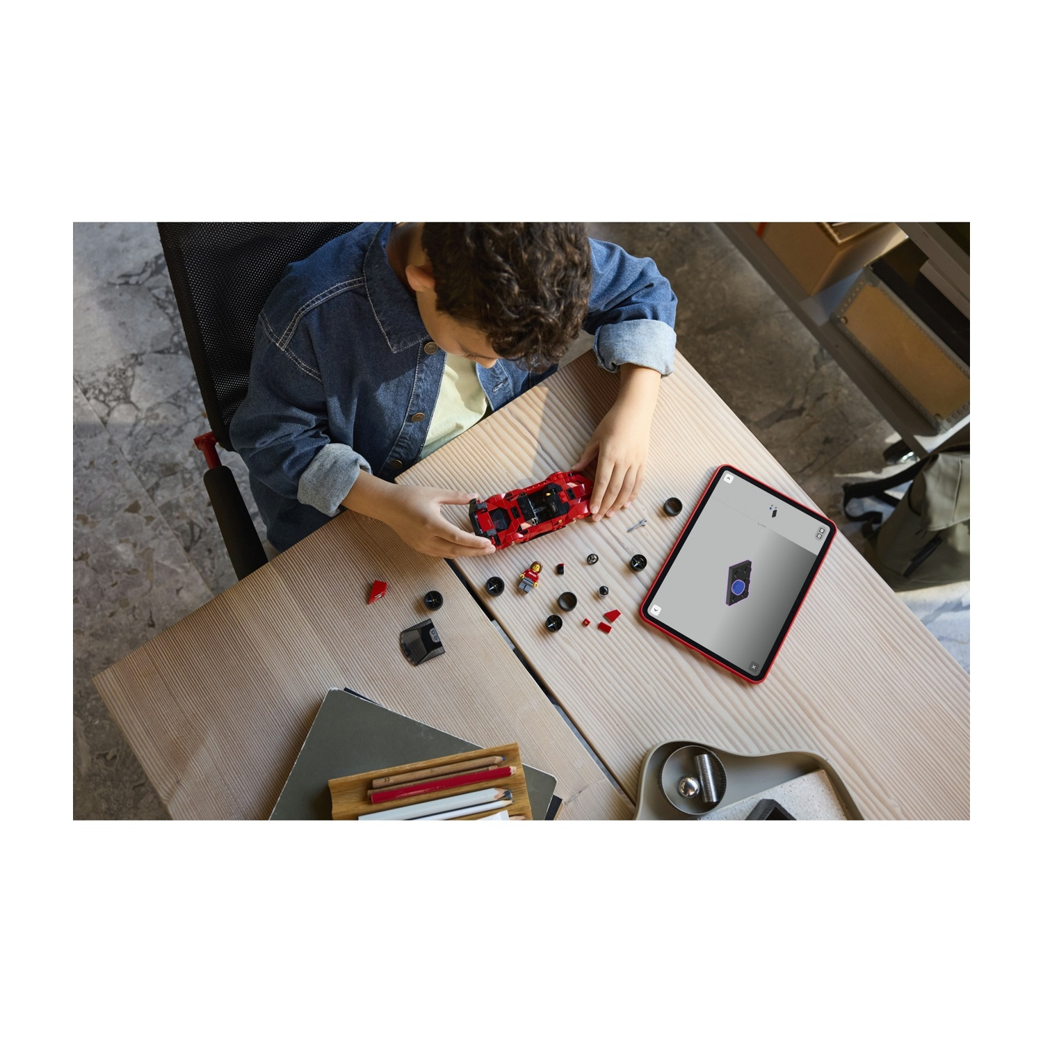Un bambino costruisce un modellino di auto rossa su un tavolo di legno. Un tablet con le istruzioni dell'auto è vicino. Parti sparse e matite sono presenti.