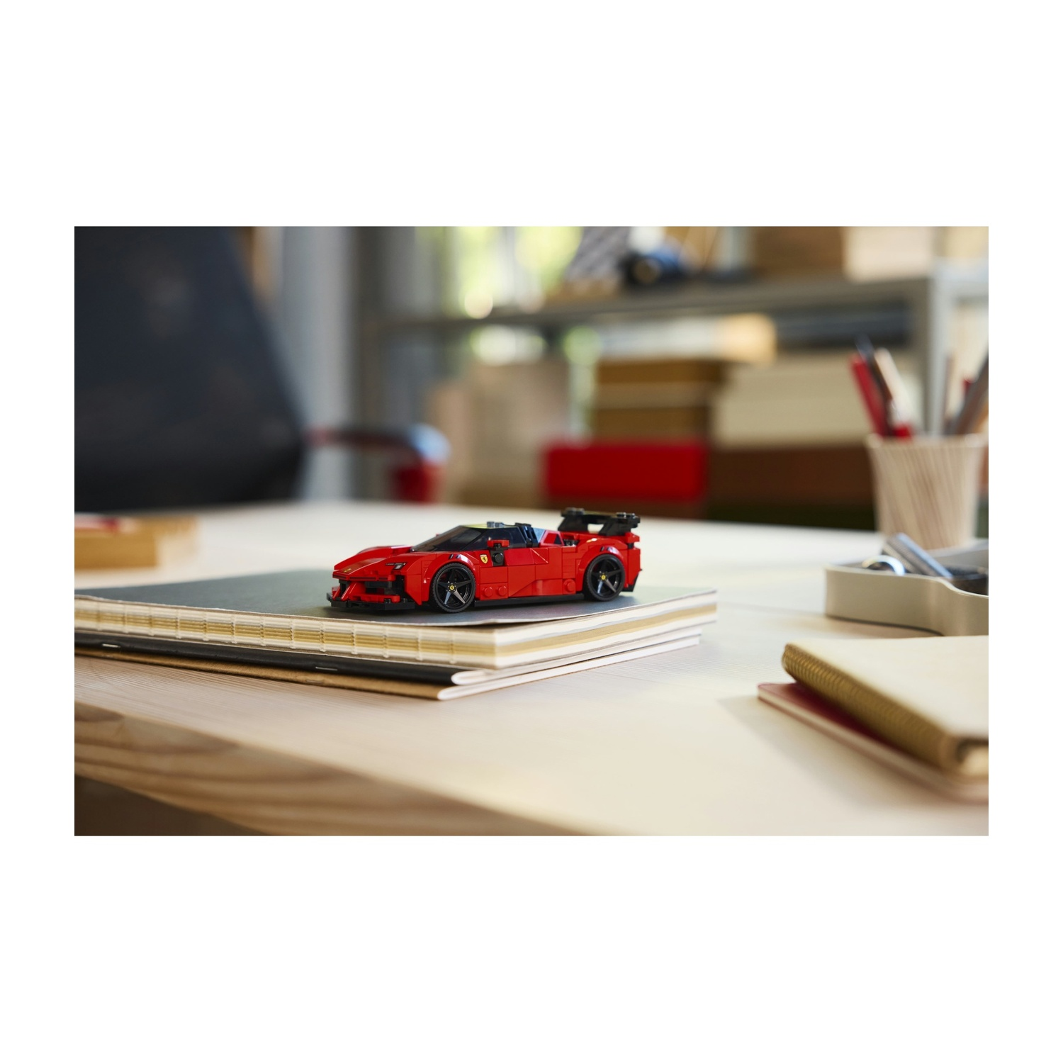 Un'auto sportiva giocattolo rossa è appoggiata su una pila di quaderni su un tavolo di legno. Lo sfondo include una scrivania, penne e una finestra.