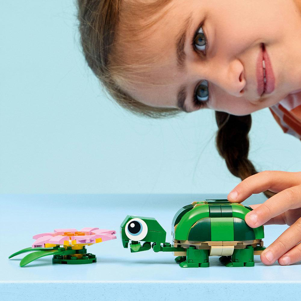 Ein Mädchen lächelt eine LEGO-Schildkröte und eine Blume an. Die Schildkröte ist grün, mit braunen und dunkelgrünen Details. Die Blume ist rosa und gelb.