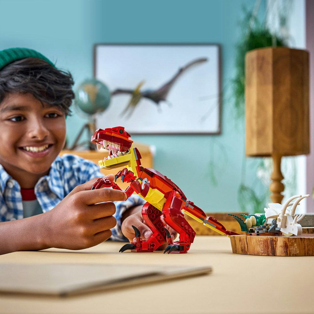 Ein lächelnder Junge spielt mit einem roten Lego-Dinosaurier. Ein Pterodaktylus-Bild und eine Lampe sind im Hintergrund.