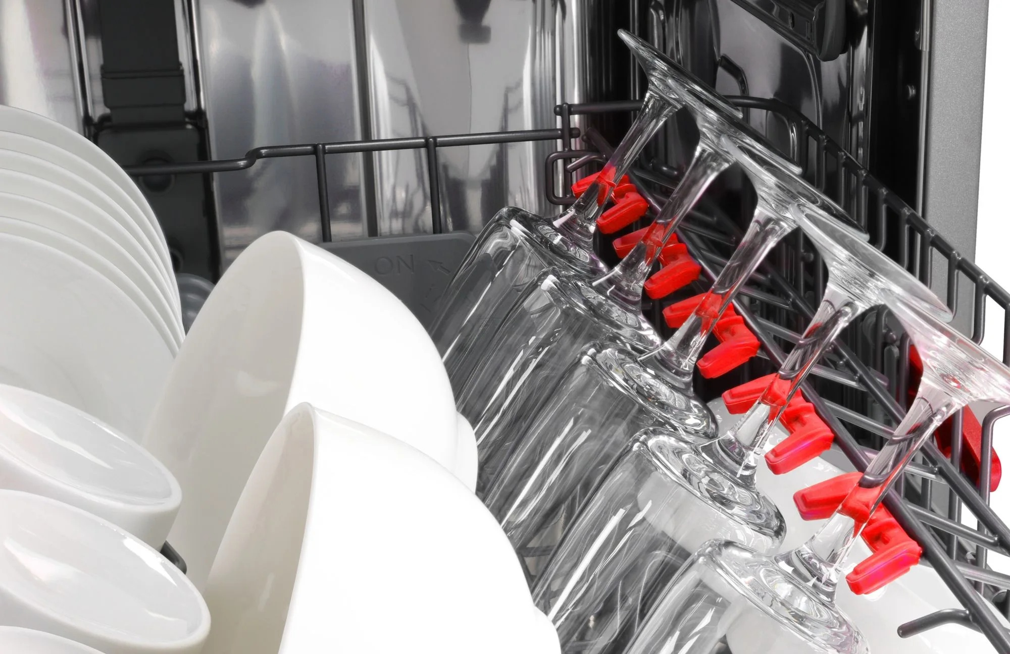 W zmywarce załadowano czyste, białe naczynia i przezroczyste szklanki. Czerwone klipsy zabezpieczają szklanki w stojaku.