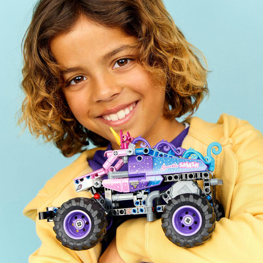 Ein lächelndes Kind hält einen Spielzeug-Monstertruck mit Einhorn-Design. Der Truck ist lila, rosa und blau. Das Kind trägt eine gelbe Jacke.