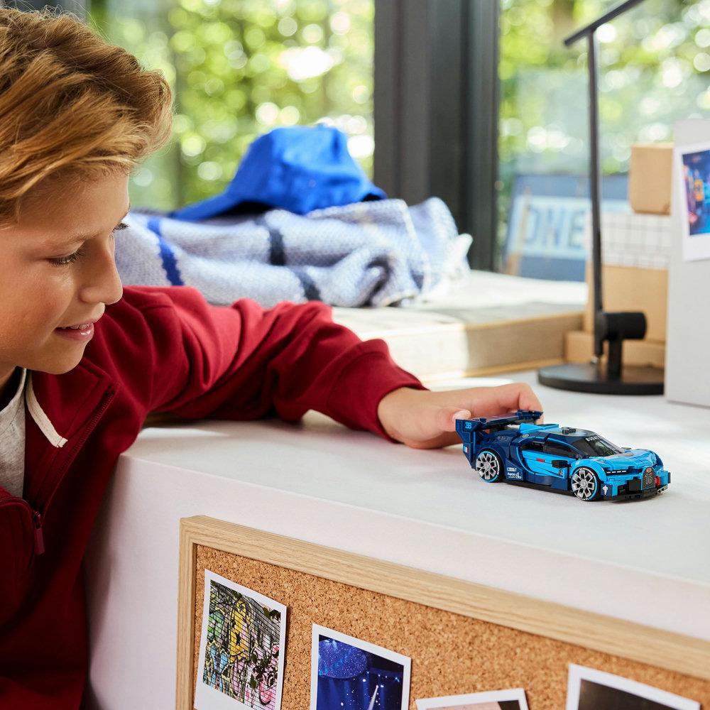Ein Junge in roter Jacke betrachtet ein blaues Spielzeugauto. Das Auto steht auf einer weißen Oberfläche. Korkwand mit Bildern im Vordergrund.