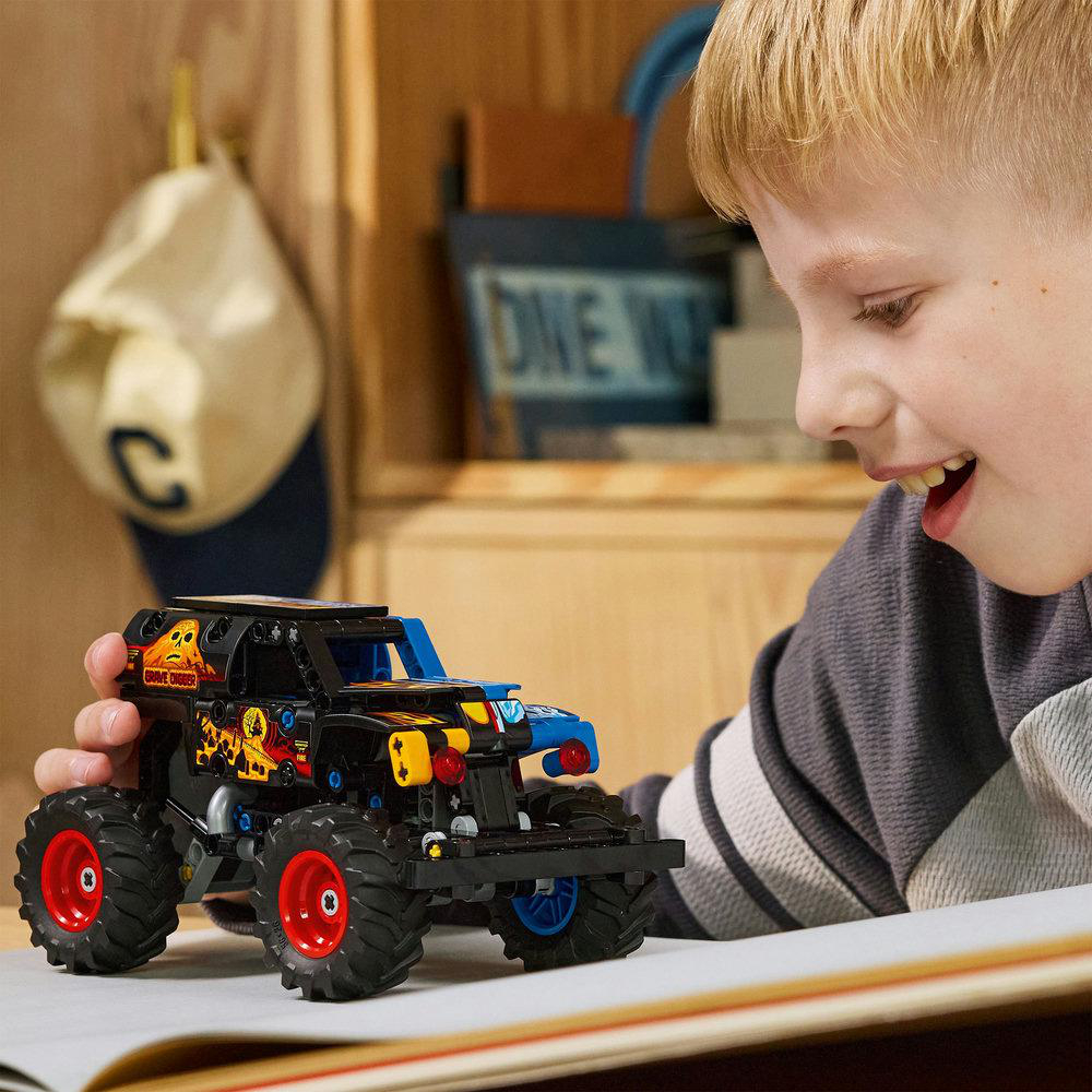 Ein Junge lächelt ein schwarzes Monstertruck-Spielzeug mit roten Rädern an. Der Truck steht auf einer weißen Oberfläche. Im Hintergrund hängt eine Mütze.