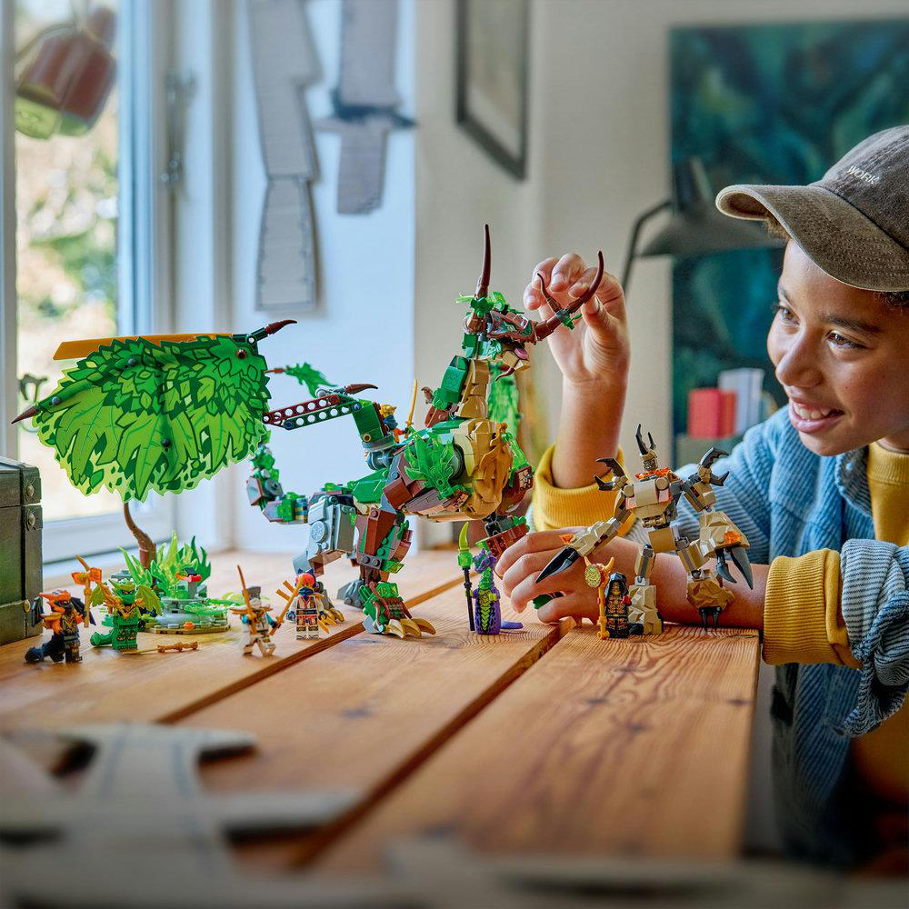 Ein Kind spielt mit einem grünen Drachen-Lego-Set. Andere Lego-Figuren stehen auf dem Holztisch. Das Kind lächelt beim Zusammenbau des Sets.