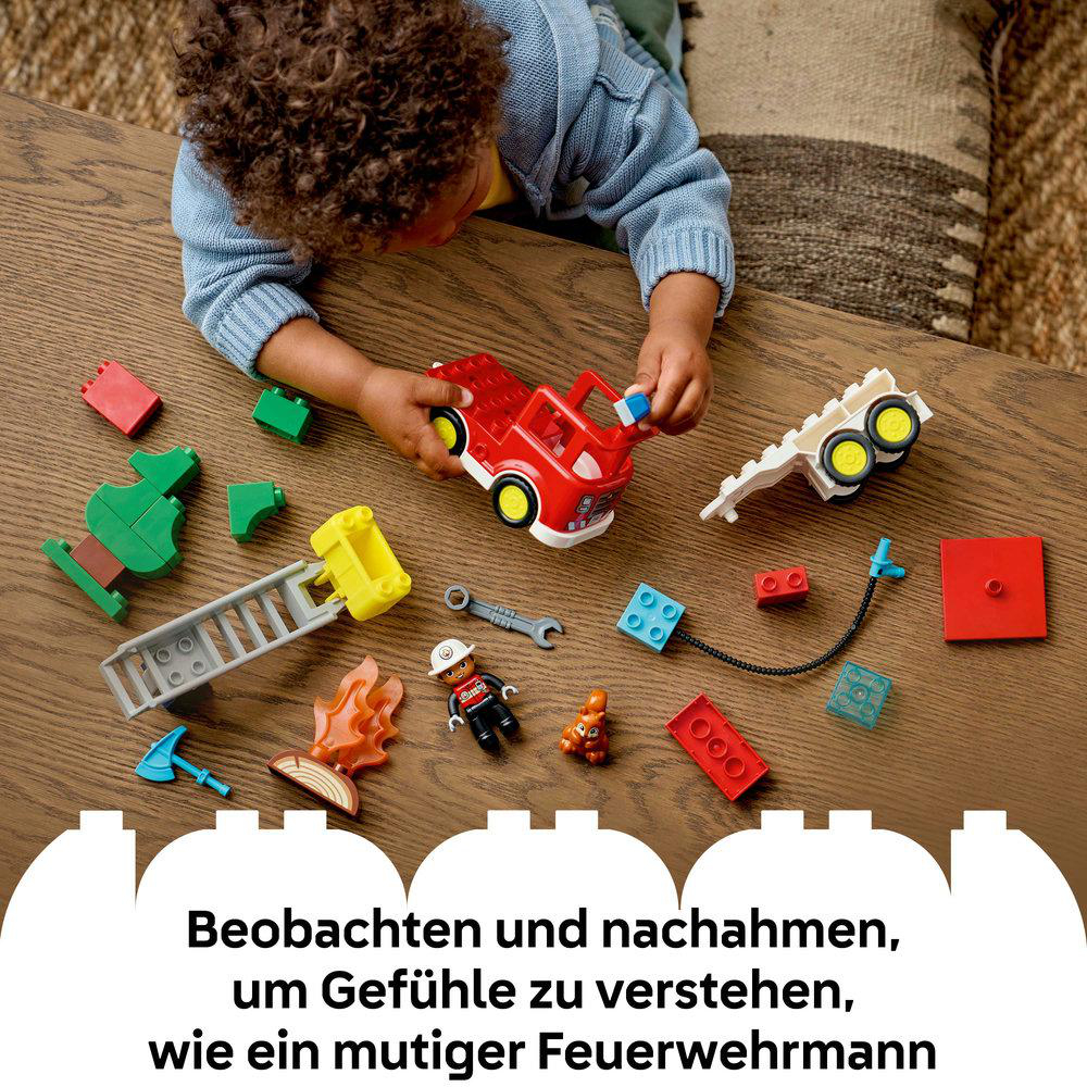 Ein Kind spielt mit einem roten Feuerwehrauto und anderen bunten Blöcken auf einem Holztisch. Die Blöcke bilden eine Feuerwehrszene.