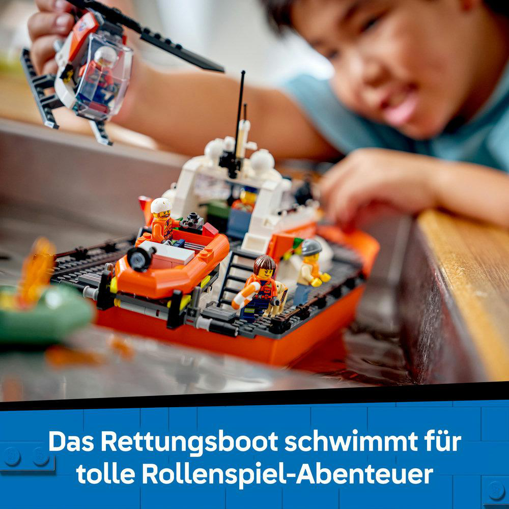 Ein Kind spielt mit einem Lego-Rettungsboot und -Hubschrauber im Wasser. Das Boot ist orange und weiß, mit Lego-Figuren. Ein Junge schaut zu.