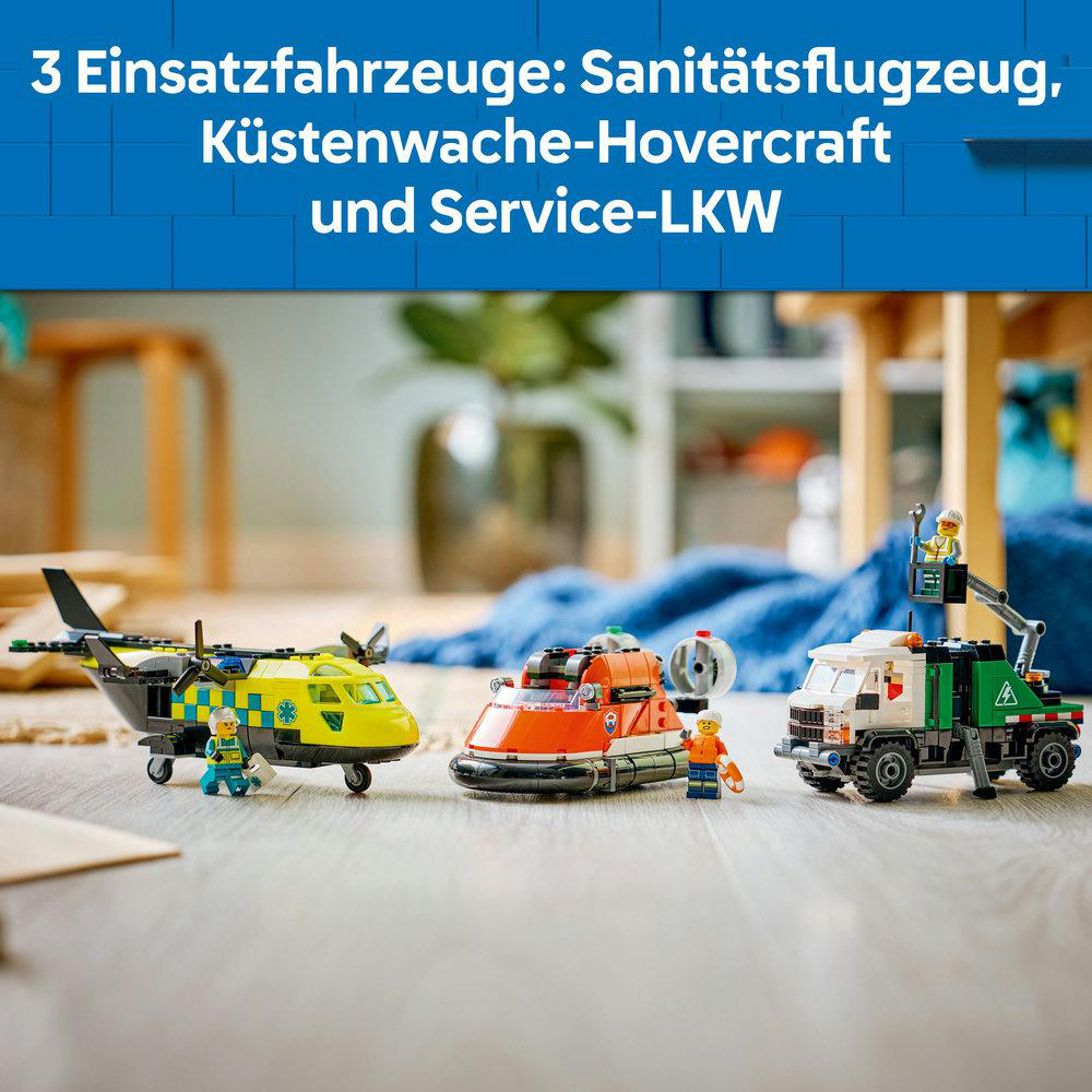 Drei Spielzeug-Notfallfahrzeuge: ein gelb-schwarzer Hubschrauber, ein orangefarbenes Hovercraft und ein weiß-grüner Lastwagen auf Holzboden.