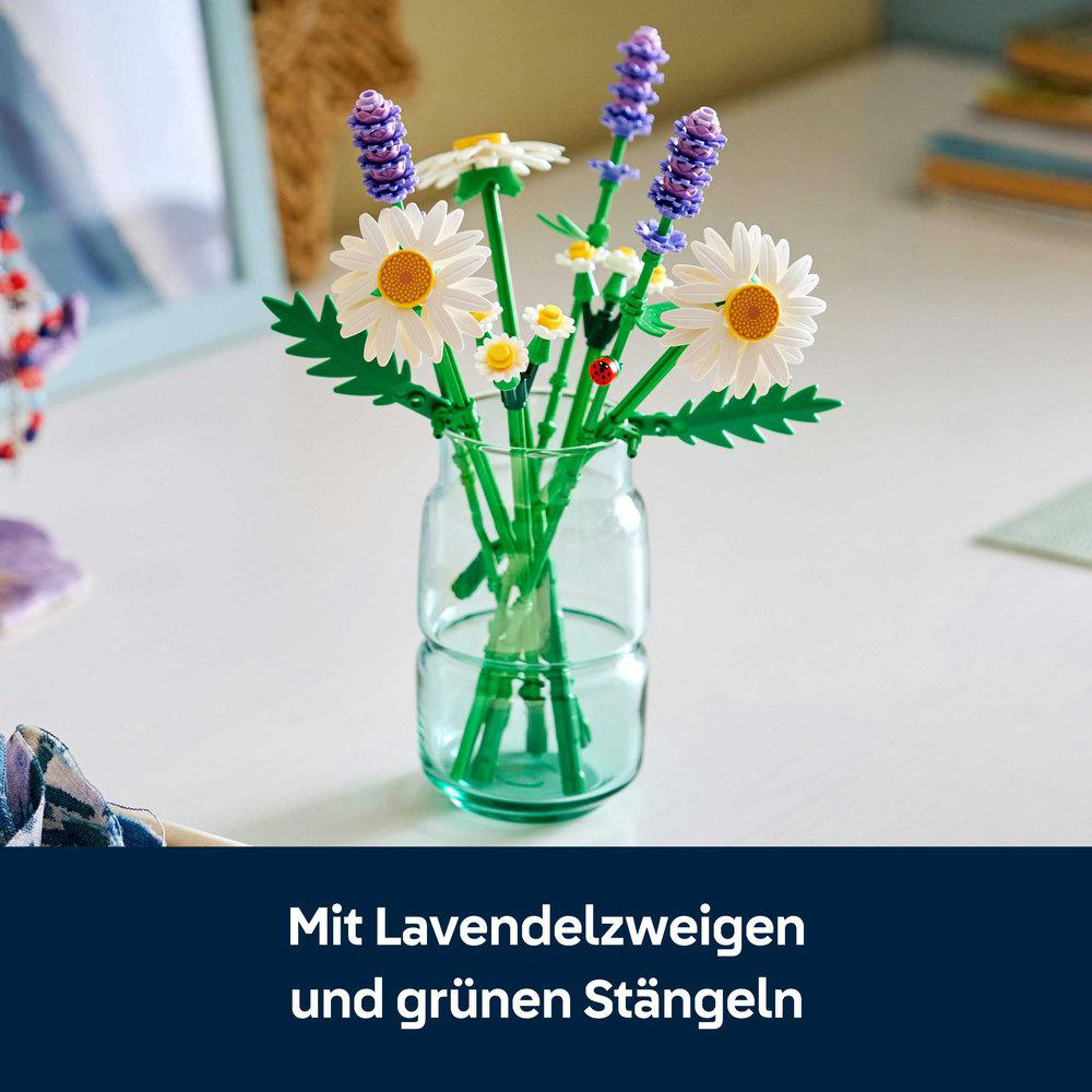 Eine Glasvase mit Lego-Gänseblümchen und Lavendelblüten. Ein Marienkäfer sitzt auf einem grünen Stiel. Der Hintergrund ist weiß.